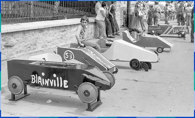 Une voiture de course (boîte à savon) est déposée sur des blocs. Un enfant est assis sur la deuxième boîte à savon en partant de la gauche. Une foule de gens se trouve à l'arrière-plan à droite.e.