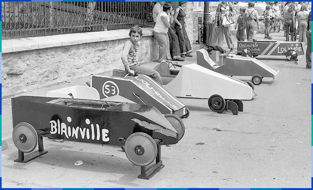Une voiture de course (boîte à savon) est déposée sur des blocs. Un enfant est assis sur la deuxième boîte à savon en partant de la gauche. Une foule de gens se trouve à l'arrière-plan à droite.e.