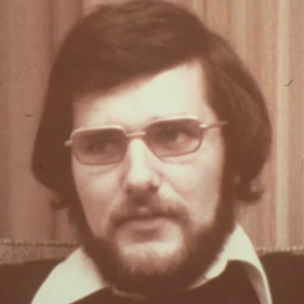 Portrait photographique d'un homme portant des lunettes et arborant une barbe et des cheveux mi-longs. Un col de chemise est visible par-dessus son chandail.