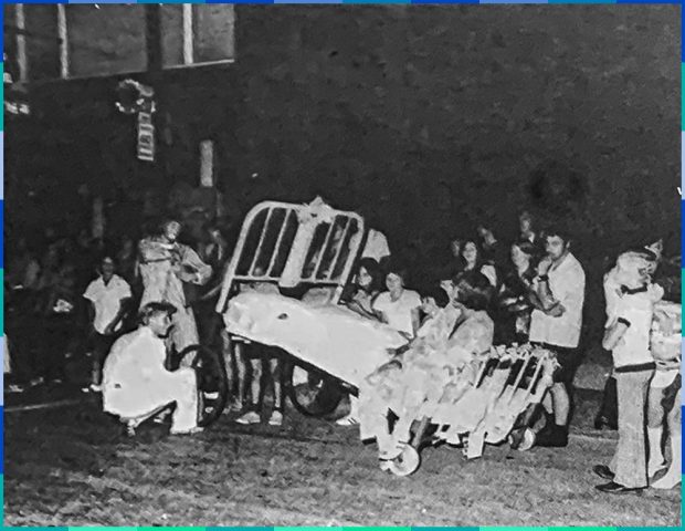 Au centre de la photographie en noir et blanc, se trouve un lit d'hôpital à barreaux métalliques sur roulettes. Une personne y est assise tandis qu'une autre semble ajuster une roue. Une petite foule les regarde.