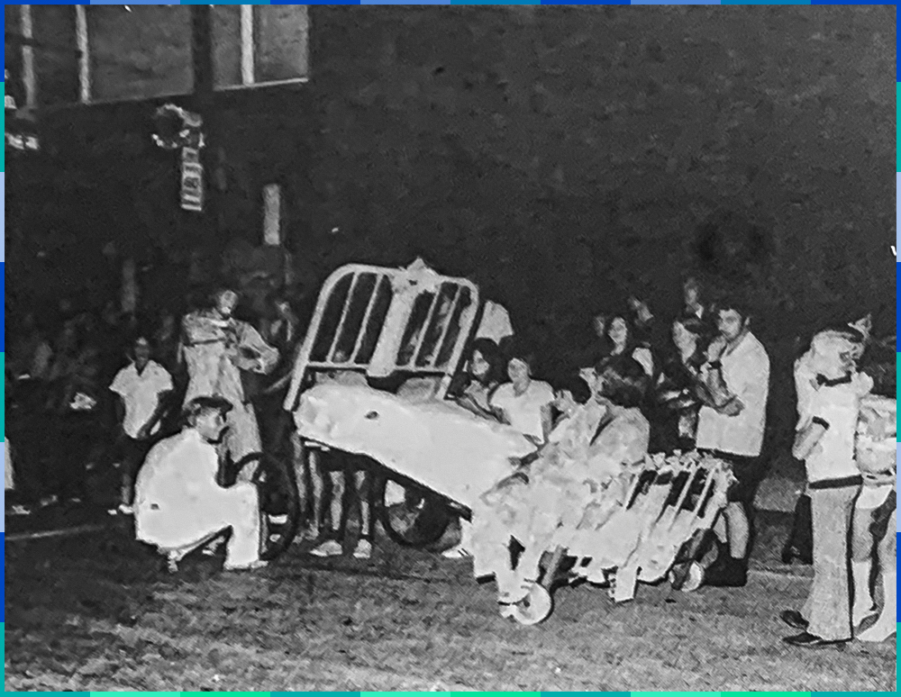 Au centre de la photographie en noir et blanc, se trouve un lit d'hôpital à barreaux métalliques sur roulettes. Une personne y est assise tandis qu'une autre semble ajuster une roue. Une petite foule les regarde.