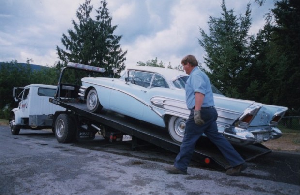 Voiture de collection bleue et blanche chargée sur une remorque attachée à une remorqueuse. Homme marche devant.