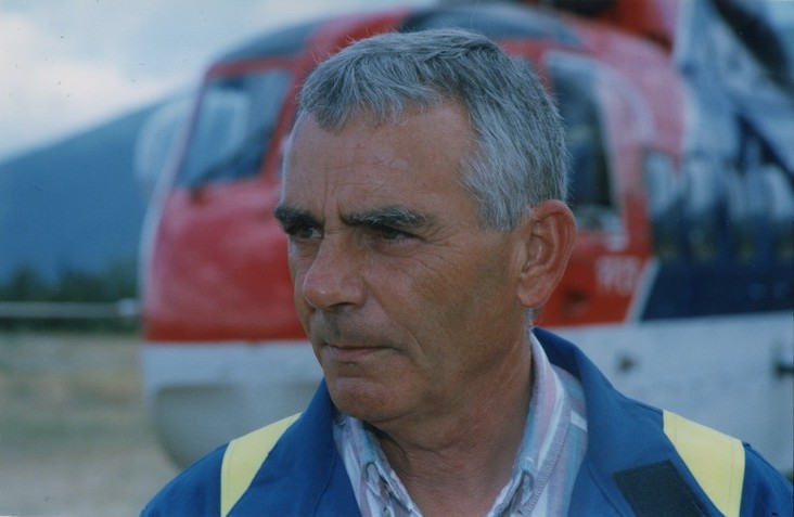 Homme au regard sérieux vêtu d'une combinaison. Hélicoptère en arrière-plan.