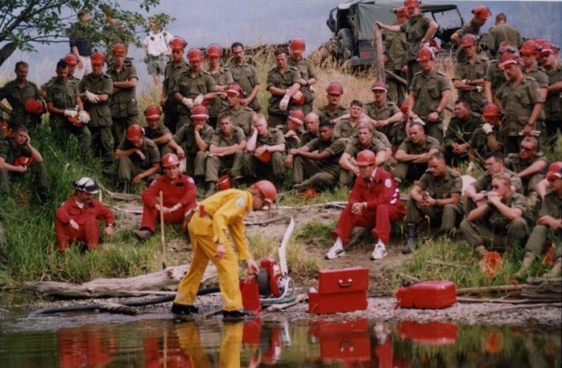 Groupe de soldats et de personnel du ministère des Forêts regardent un homme en combinaison jaune travailler avec une pompe à eau. La plupart portent des casques de sécurité. Eau au premier plan.