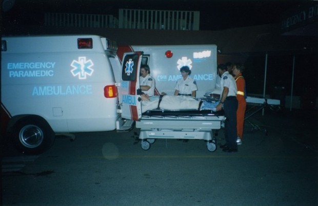 Trois ambulanciers metten un homme sur une civière dans l'ambulance. Ouvrier en uniforme orange avec bandes réfléchissantes regarde.