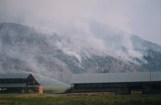 Incendie sur les collines derrière une ferme, granges devant. Eau aspergeant les champs.