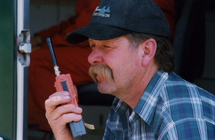 Homme vêtu d'une chemise bleue et blanche et d'une casquette bleue parle dans un radiotéléphone.