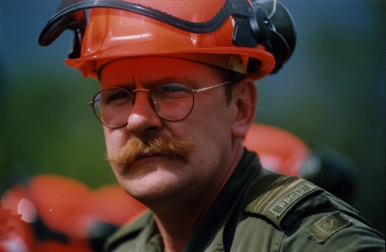 Homme moustachu avec lunettes, en uniforme vert coiffé d'un casque de sécurité, regarde la caméra en clignant des yeux.