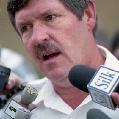 Homme moustachu, vêtu d'une chemise blanche, parle dans des microphones.