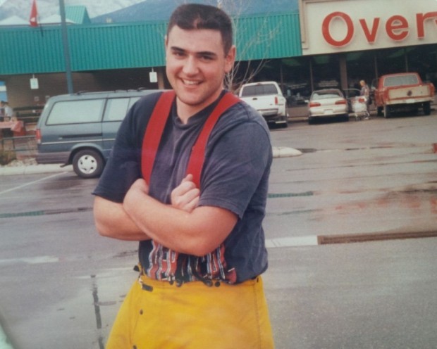 Debout dans un stationnement mouillé, jeune homme souriant portant des pantalons imperméables jaunes et des bretelles.