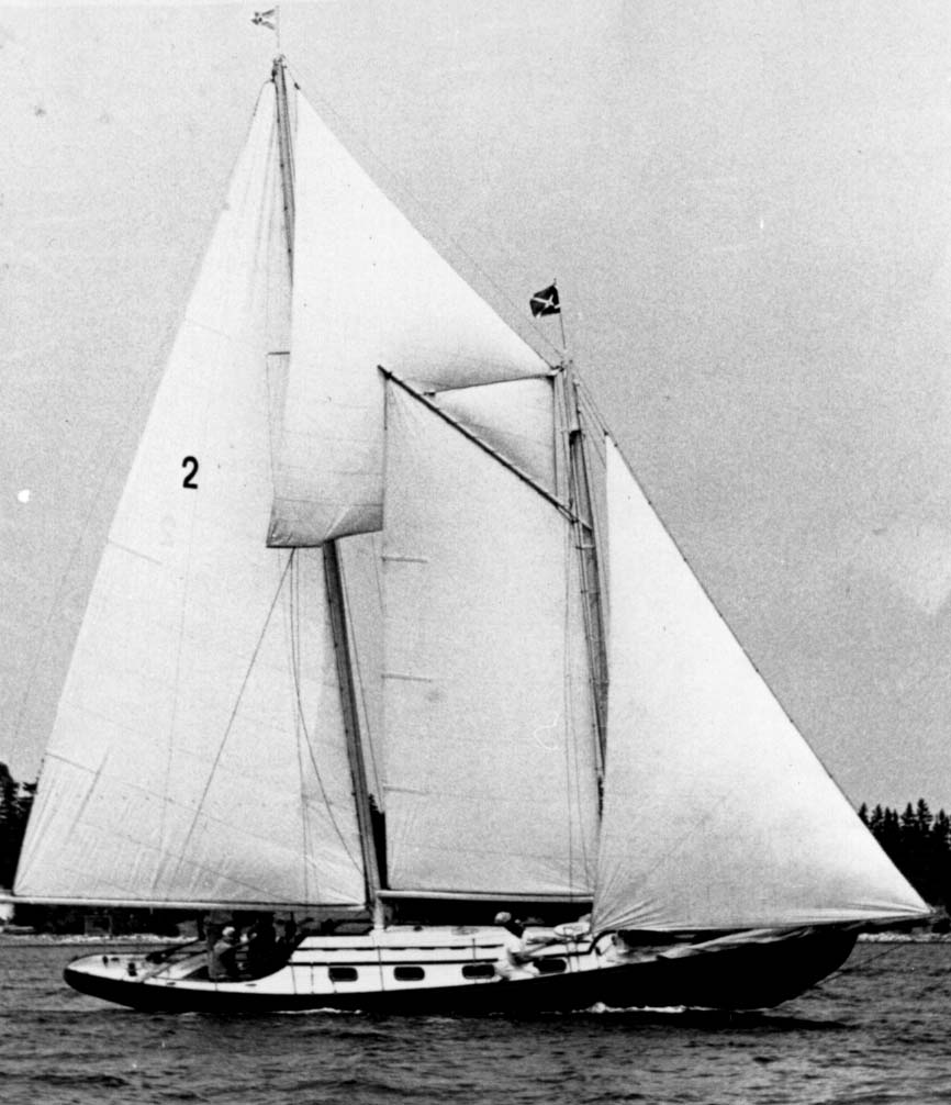 Une photo en noir et blanc de la goélette «Airlie » toutes voiles dehors dans le port. Le « Airlie » est noir avec une cabine blanche, des voile blanches et le numéro d’identification 2 sur la voile principale parmi les cinq voiles utilisées dans la photo. Des goélettes comme celle-ci étaient utilisées pour la pêche. Elles ont rapidement été adaptées pour les nouveaux moteurs «make and break ». De nos jours elles sont principalement utilisées comme bateaux de plaisance.