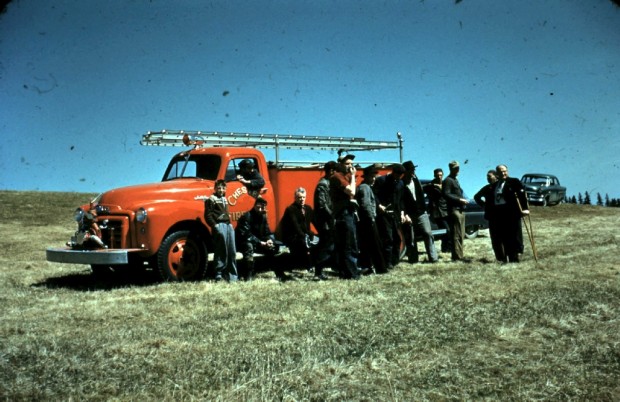 Une photo en couleur montrant, vers 1937, le premier camion de pompiers rouge avec les pompiers se tenant devant lui. Le camion a une échelle sur le toit et une pompe à eau sur le pare-choc avant.