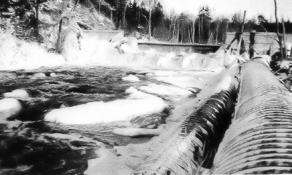 Une photo en noir et blanc montrant le barrage sur la East River avec les vannes fermées, l’eau tourbillonnant au pied du barrage et ensuite s’écoulant le long de la rivière. Sur la rive droite, deux grosses canalisations acheminent l’eau à la centrale électrique.