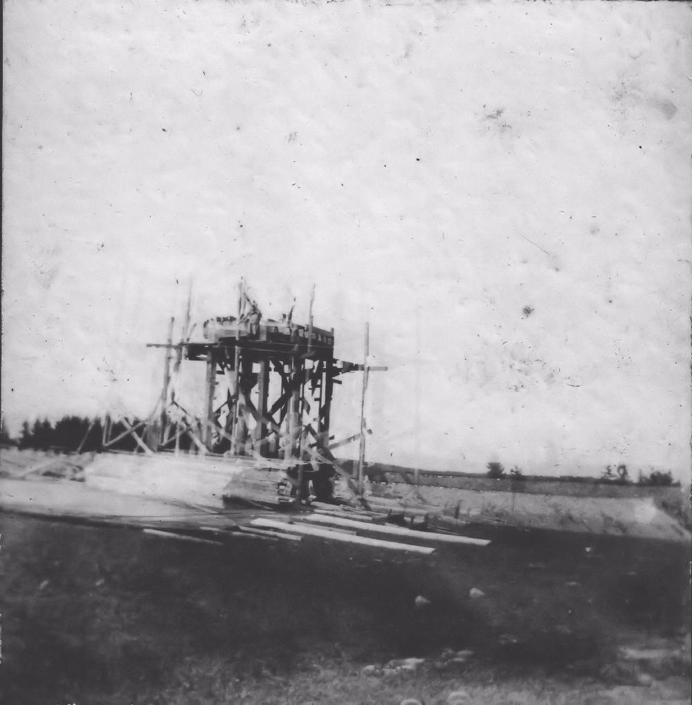 Une photo noir et blanc montrant la charpente du château d’eau en construction. On peut voir des piles de bois au premier plan et le talus de la voie ferrée de la Halifax and Southwestern Railway à l’arrière-plan. Le château d’eau devait fournir l’eau pour le système de canalisations d’eau du village et pour les trains.