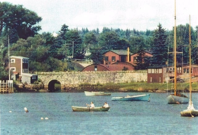 Une photo couleur avec des bateaux au premier plan   prise   de Mill Cove en direction de Hawboldt Gas Engines. On peut voir le vieux pont de pierre, le bâtiment rond et rouge de la fonderie, l’atelier et le bureau principal. Un bâtiment à gauche du pont servait d’entrepôt. Les bâtiments rouges de l’autre côté du pont étaient une menuiserie. Le ruisseau prenait sa source dans le lac Stanford. Il permettait d’alimenter en électricité la fonderie et fournissait de l’eau au système de canalisations d’eau du village. Il se jetait dans Mill Cove sous le vieux pont de pierre.