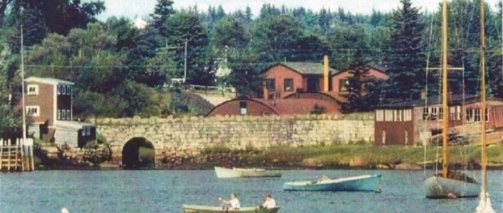 Une photo couleur avec des bateaux au premier plan   prise   de Mill Cove en direction de Hawboldt Gas Engines. On peut voir le vieux pont de pierre, le bâtiment rond et rouge de la fonderie, l’atelier et le bureau principal. Un bâtiment à gauche du pont servait d’entrepôt. Les bâtiments rouges de l’autre côté du pont étaient une menuiserie. Le ruisseau prenait sa source dans le lac Stanford. Il permettait d’alimenter en électricité la fonderie et fournissait de l’eau au système de canalisations d’eau du village. Il se jetait dans Mill Cove sous le vieux pont de pierre.