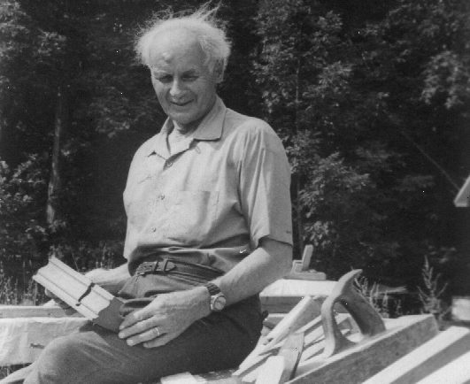 Photographie noir et blanc de François-Xavier Lachance, âgé, assis à l’extérieur sur une pile de matériaux, tenant une moulure en bois.