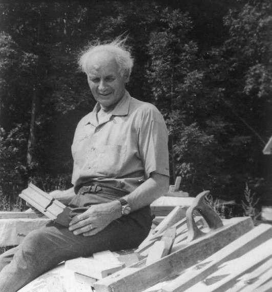 Photographie noir et blanc de François-Xavier Lachance, âgé, assis à l’extérieur sur une pile de matériaux, tenant une moulure en bois.