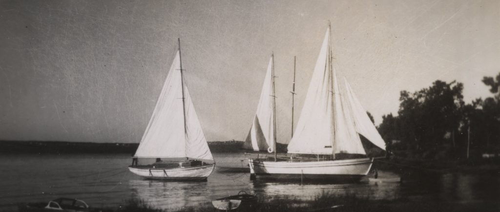 Photographie noir et blanc de trois yachts blancs, aux voiles hissées, ancrés près de la rive.