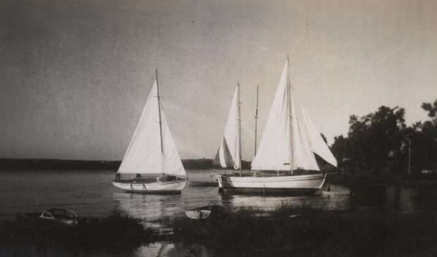 Photographie noir et blanc de trois yachts blancs, aux voiles hissées, ancrés près de la rive.