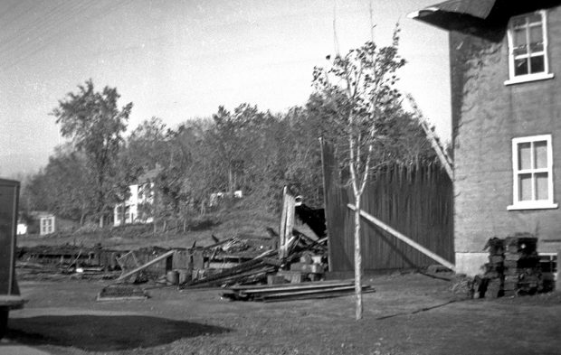 Photographie noir et blanc montrant la fondation d’un bâtiment, un mur de bois brûlé presque en entier et des planches brûlées au sol. Une résidence en pierre, dont le mur gauche et le toit de la maison sont noircis par le feu, est partiellement visible, à droite de l’image.