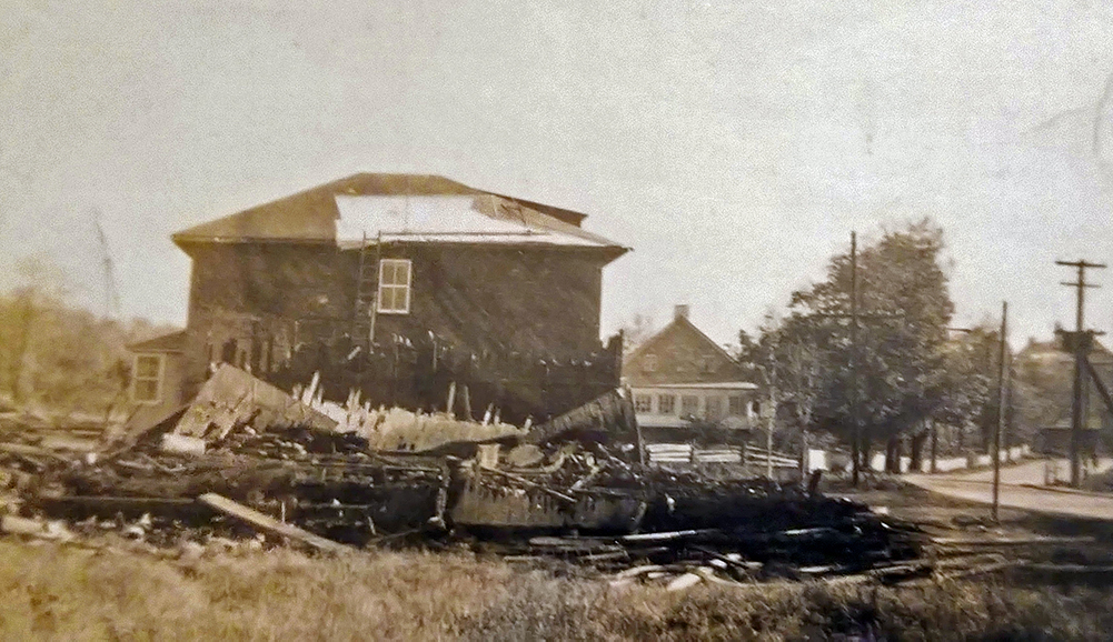 Photographie sépia montrant un amas de planches brûlées à l’avant-plan. Une résidence à deux étages endommagée par le feu est visible derrière les vestiges. 