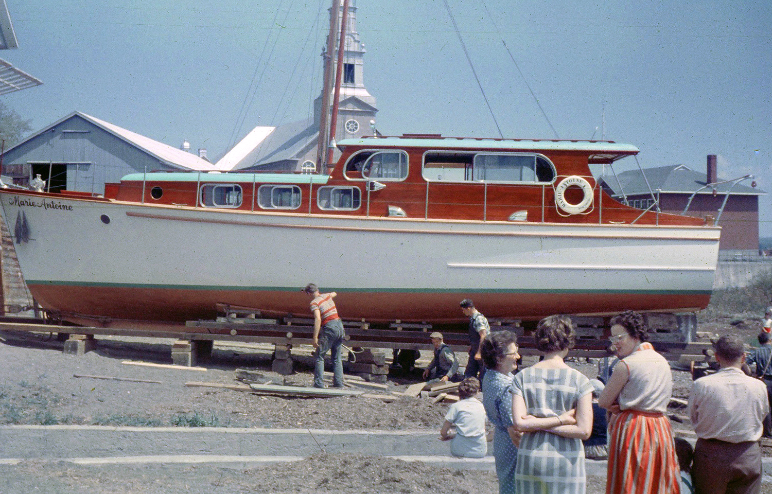 Photographie couleur où un bateau de plaisance en bois peint en blanc, dont la cabine est de couleur acajou, est installé sur des blocs et des planches. Le bateau est montré de profil, avec une église et un hangar à l’arrière-plan. L’inscription « Marie-Antoine » se trouve à l’avant du bateau. Trois hommes, travaillent sur la structure sous l’embarcation. Trois femmes discutent à l’avant-plan. Un homme observe la scène.