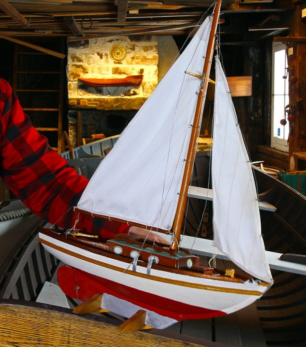 Photographie couleur de la maquette d’un voilier en bois, vue de profil. Le bateau est peint en blanc, rouge et brun. Les deux voiles blanches sont hissées. 