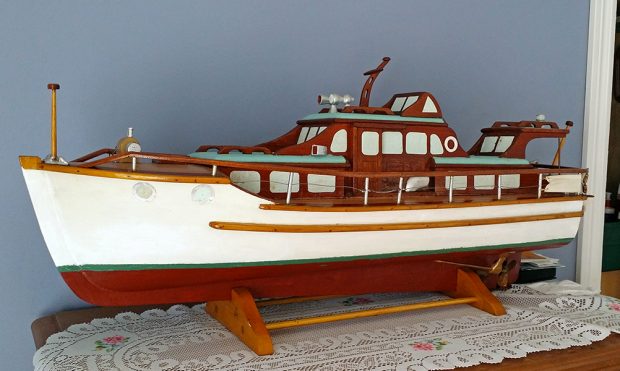 Photographie couleur de la maquette d’un yacht en bois, vue de profil. Le bateau est peint en blanc, rouge, vert et brun. L’objet est exposé sur un petit socle en bois, sur le dessus d’un meuble.