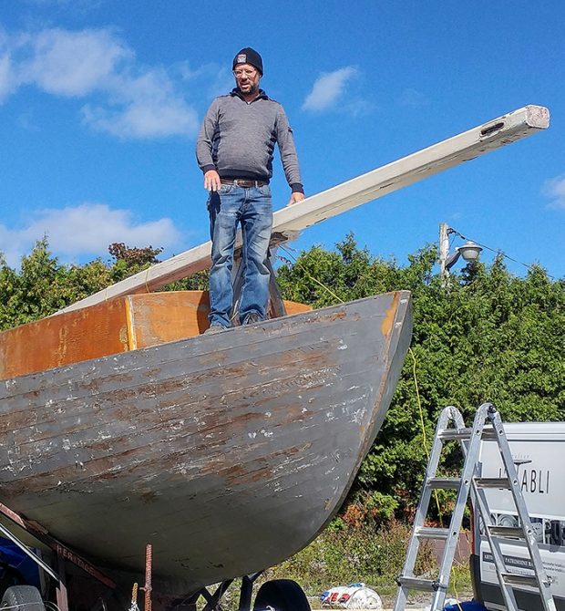 Photographie couleur en contre-plongée montrant le devant d’un bateau de bois peint en gris, usé, sur lequel se tient un homme debout (Jean-François Lachance), appuyé sur le mat couché du bateau posé sur une remorque. 