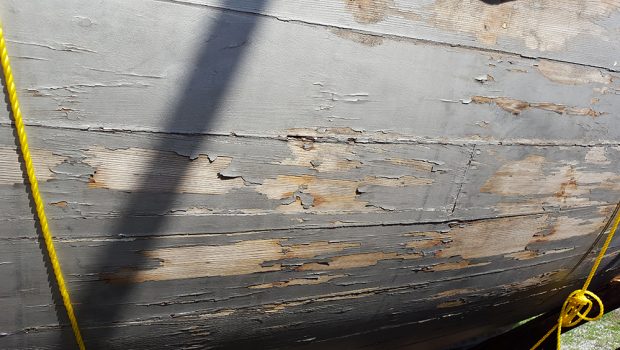 Photographie couleur montrant le détail de la coque d’un bateau de bois peint en gris, dont la peinture est écaillée. 