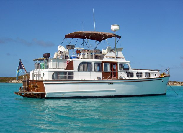 Photographie couleur d’un yacht blanc, de profil, sur une mer bleu azur. Le pont supérieur est aménagé en terrasse avec barbecue et abri en toile.