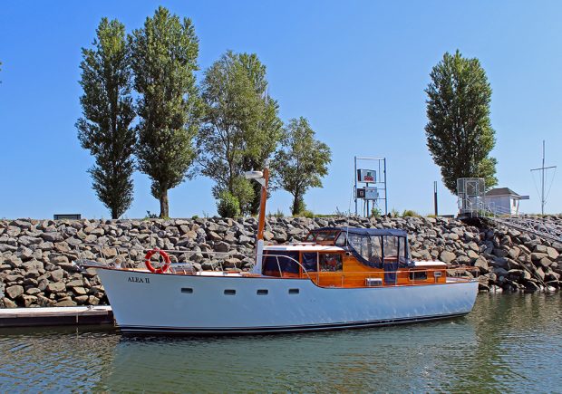 Photographie couleur d’un voilier blanc, de profil, portant l’inscription «ALEA II», amarré à un ponton de bois devant un brise-lame rocheux. Une descente pour piétons se trouve sur la droite de l’image. 