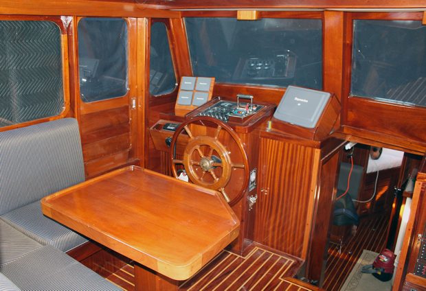 Photographie couleur de l’intérieur d’une cabine, dont les surfaces sont en boiserie. Au centre se trouve la roue, devant les instruments de navigation. Une table en bois entourée en partie d’une banquette est à l’avant-plan. 