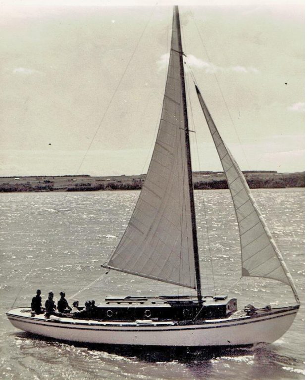 Photographie d’un voilier blanc à cabine foncée, qui navigue voiles déployées, sous le soleil. Six personnes sont à bord. Des champs sont visibles à l’arrière-plan.