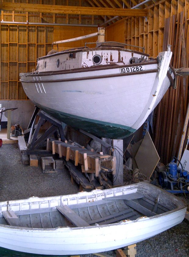 Photographie couleur d’un bateau de bois blanc, brun et vert, posé sur un tréteau à l’intérieur d’un bâtiment. 