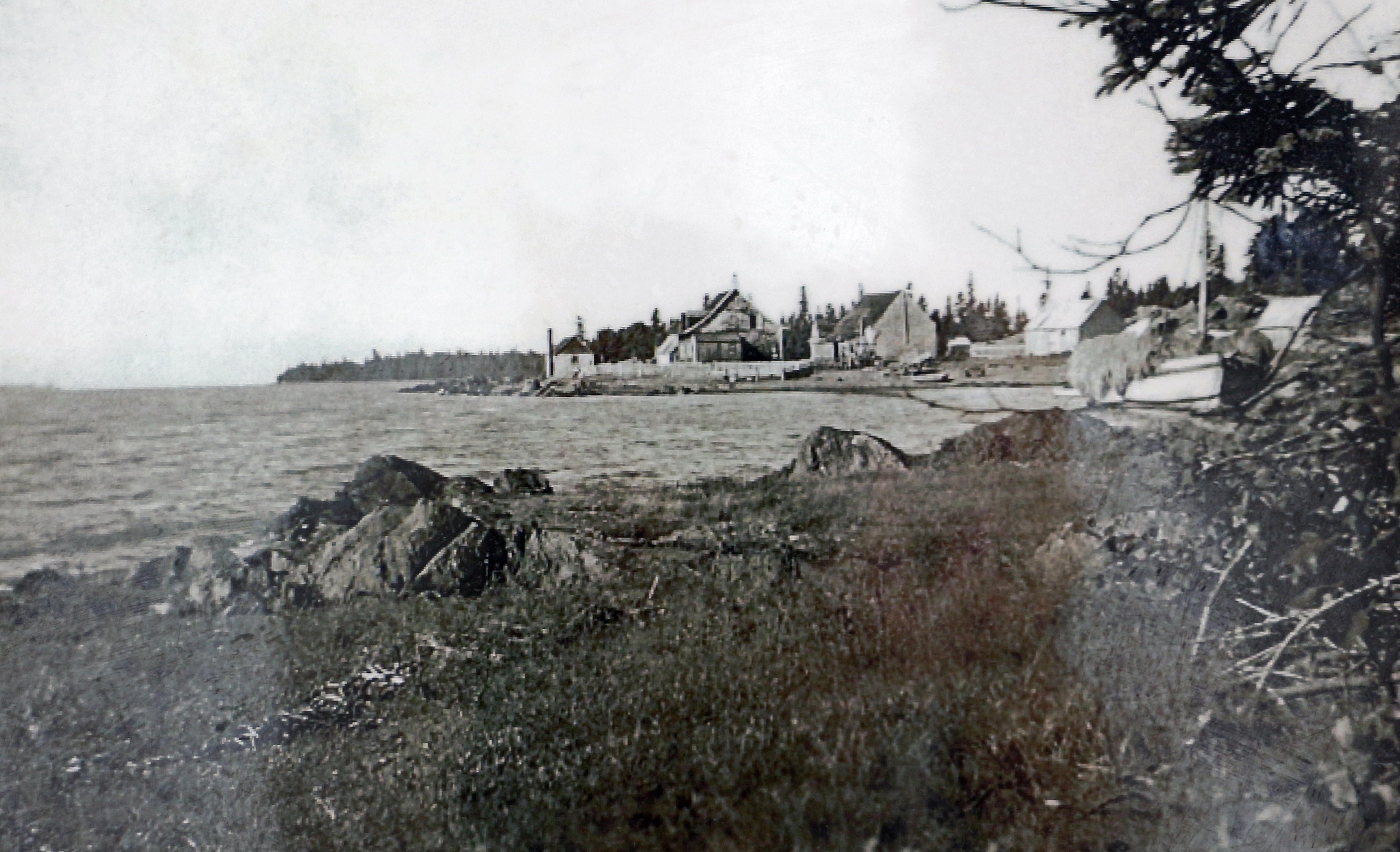 Photographie noir et blanc qui présente quatre bâtiments en bois, à l'arrière-plan. Devant se trouve une baie où une petite embarcation à mat est accostée sur la berge rocheuse.