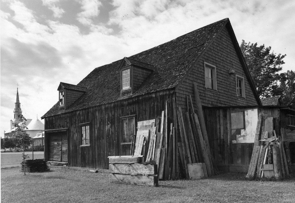 Photographie noir et blanc d'un bâtiment en bois et bardeaux de cèdre, muni de deux lucarnes. Plusieurs planches et matériaux y sont adossés. L'église de Saint-Laurent est visible à l'arrière-plan, à gauche.