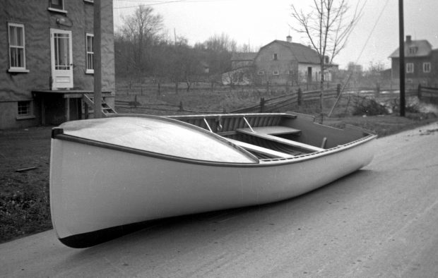 Photographie noir et blanc d'une grande chaloupe en bois blanche, à l'apparence neuve, posée sur la route. Deux maisons, un champ et une grange sont visibles à l'arrière-plan. 