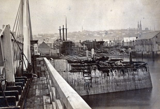 Photographie noir et blanc d’un chantier maritime. On y voit un quai à l’avant-plan, ainsi qu’un voilier à trois mats et un second sans mat au centre de l’image. D’imposants bâtiments se situent en bordure de l’eau sur la droite. La haute-ville de Québec est visible à l’arrière-plan.