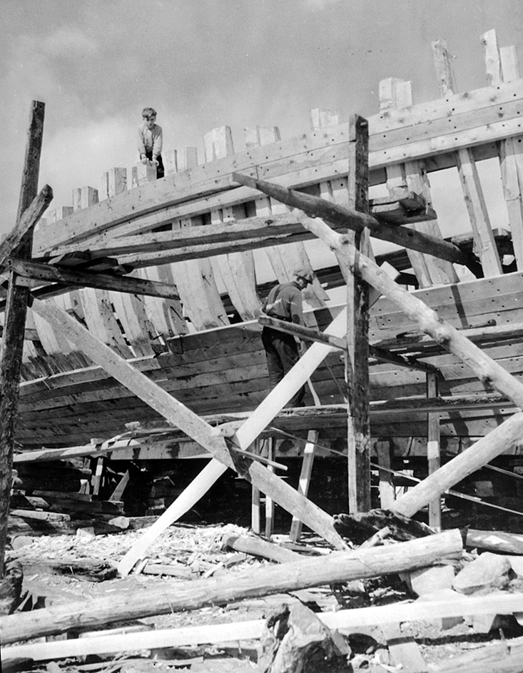 Photographie noir et blanc où un homme travaille à l’installation du revêtement d’un grand bateau, installé sur des socles. Il est debout sur un échafaudage artisanal en bois. Un jeune garçon est debout tout en haut de la structure de la cale.