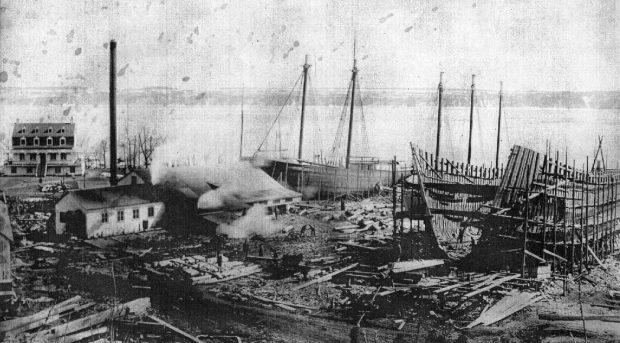 Photographie noir et blanc du chantier maritime de Saint-Laurent. La structure d’un imposant bateau se trouve sur la droite, à l’avant-plan. Deux voiliers en construction se trouvent au centre. Le fleuve Saint-Laurent occupe tout l’arrière-plan, ainsi que la côte de la rive sud.