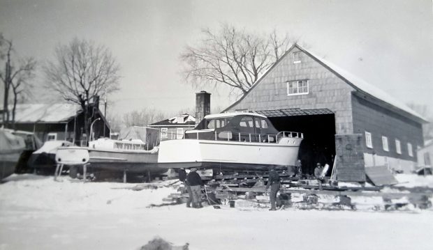 Photographie noir et blanc montrant un bateau de plaisance monté sur un socle, devant un bâtiment muni d’une grande porte, l’hiver. Quatre hommes s’activent autour du bateau, au sol. D’autres bateaux sont visibles sur la gauche.