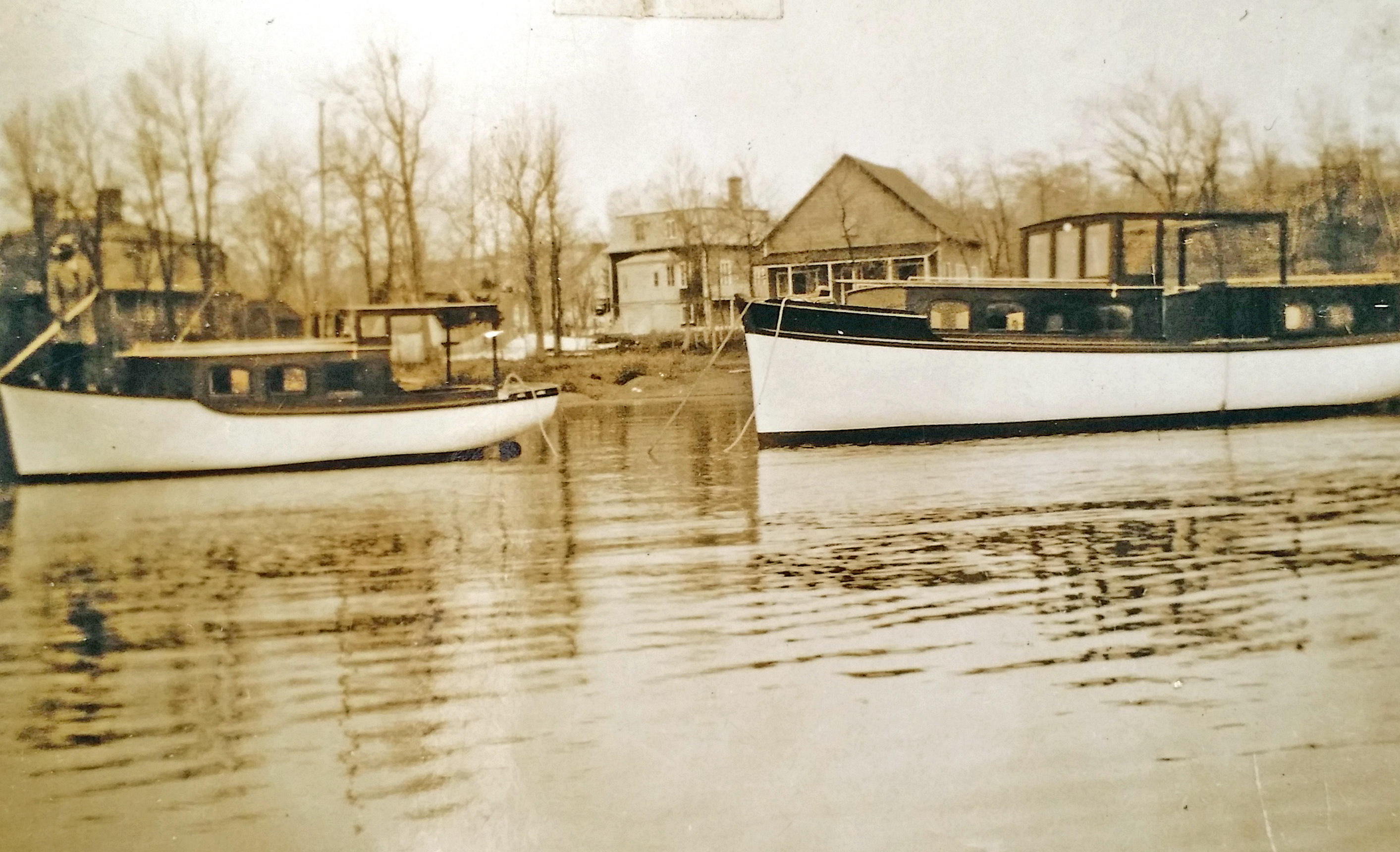 Photographie sépia de deux bateaux de bois, avec cabine, ancrés près de la rive. Des maisons et des arbres sans feuillage sont à l'arrière-plan.