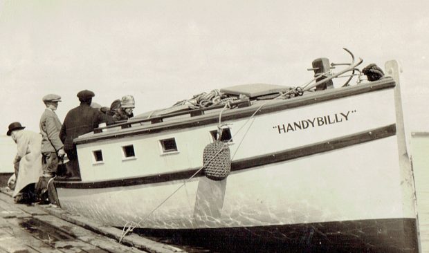 Photographie noir et blanc d’un yacht portant l’inscription « Handy Billy », accosté à un quai de bois. Une femme, deux hommes et un garçon montent à bord.