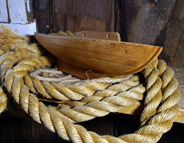 Photographie couleur d’une demi-coque en bois, vue de profil. L’objet est posé sur des cordages de bateaux.