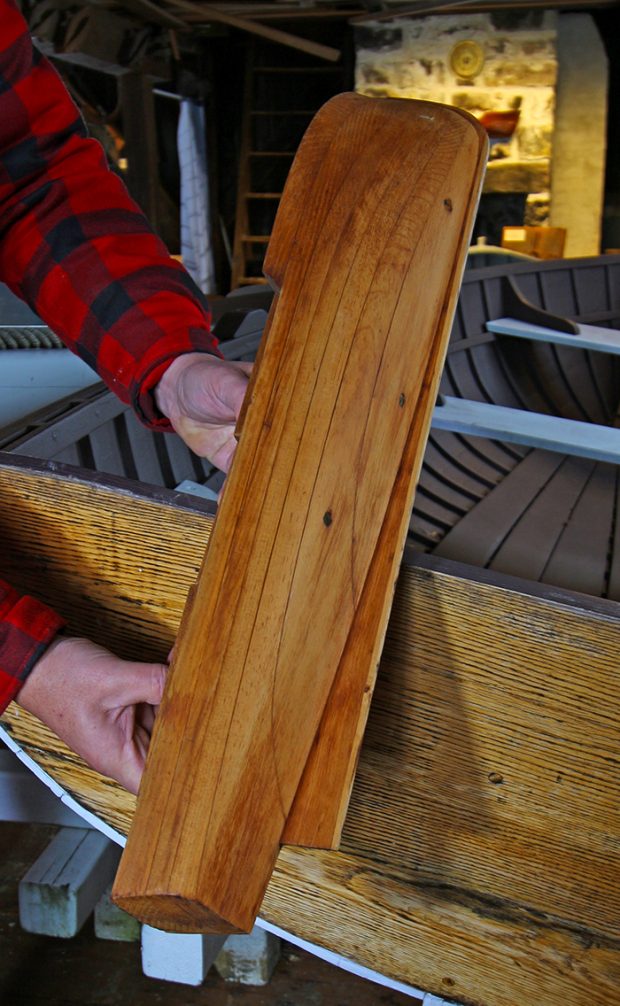 Photographie couleur d’une demi-coque en bois, présentée à la verticale, dont on voit le profil et le dessous. 
