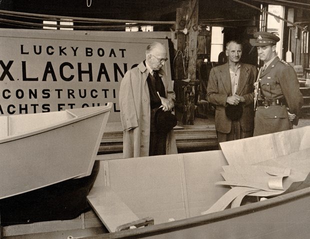 Photographie noir et blanc où trois hommes se tiennent dans une chalouperie. Une enseigne portant l’inscription « Lucky boat. FX. Lachance. Construction de yachts et chaloupes » est partiellement visible derrière eux. Les hommes de gauche (le Gouverneur général) et de droite (un militaire) regardent les embarcations. F-X Lachance est au centre, tenant un chapeau.