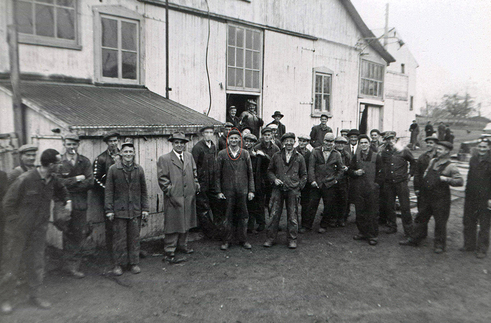 Photographie noir et blanc d’un groupe d’une trentaine d’ouvriers qui posent devant le mur latéral d’un bâtiment de planches, à l’extérieur. Les hommes sont vêtus d’une chienne de travail ou d’une salopette. Plusieurs fument et sourient en regardant la caméra. D’autres regardent vers François-Xavier Lachance, au centre. À sa droite se trouve un homme portant un manteau habillé, cravate et chapeau. 