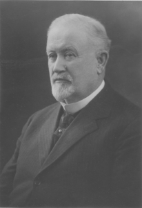 Photographie de studio d’un homme aux cheveux blanc qui porte un complet noir.